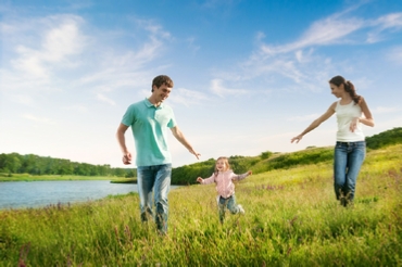 A couple run through wild grass next to a wide river, each stretching their hands out to their child running alongside between them and reaching for them both.