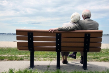 An older couple nestled together with his arm around her sit on a wooden bench overlooking a sandy beach.